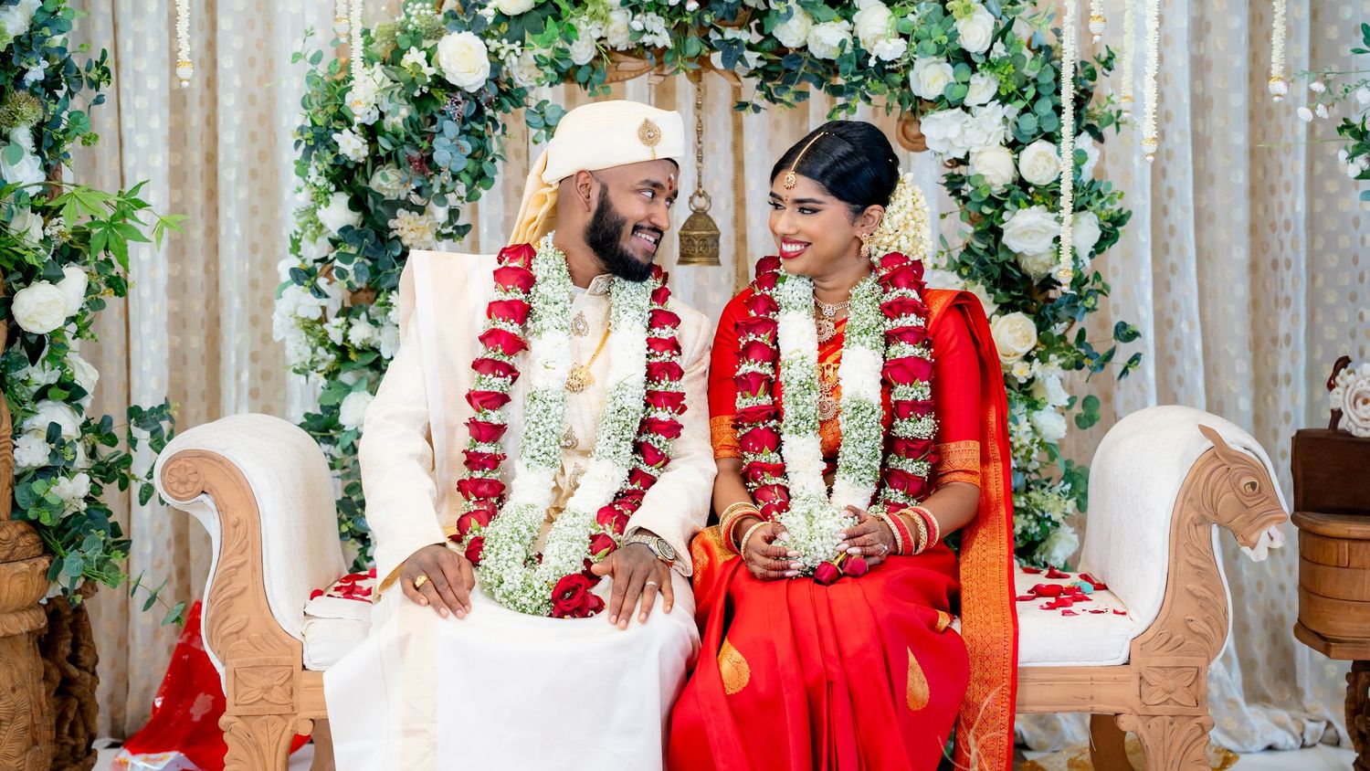 Joyful South Indian newlyweds seated together wearing red and white floral garlands on decorated wedding mandap.