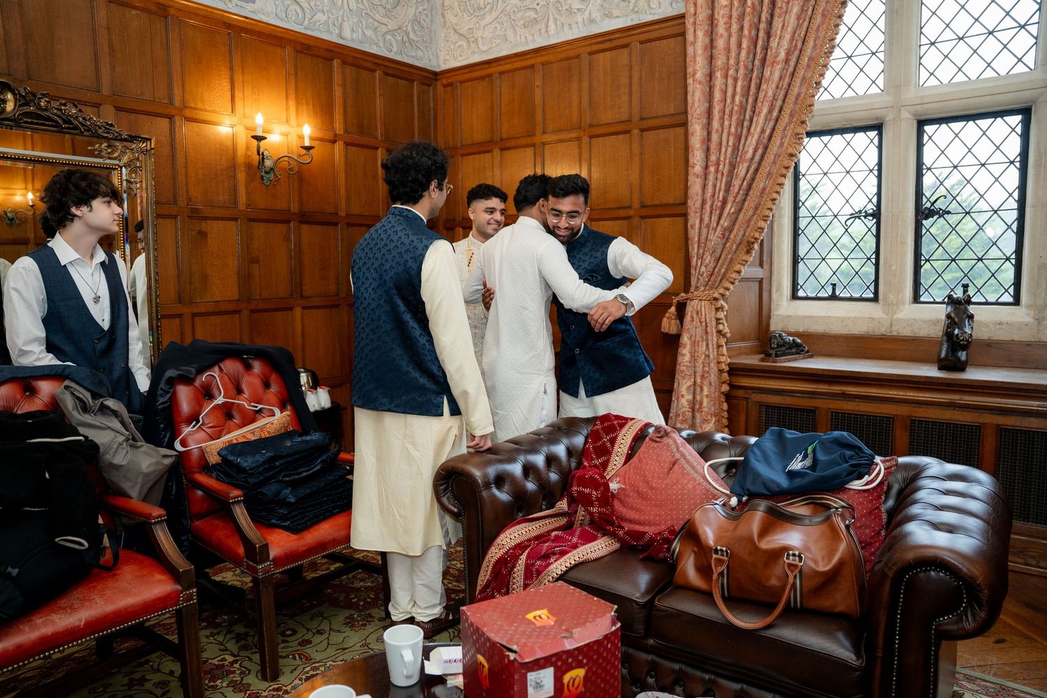 Groom in white kurta embraced by groomsmen in navy suits in wood-paneled wedding preparation room.