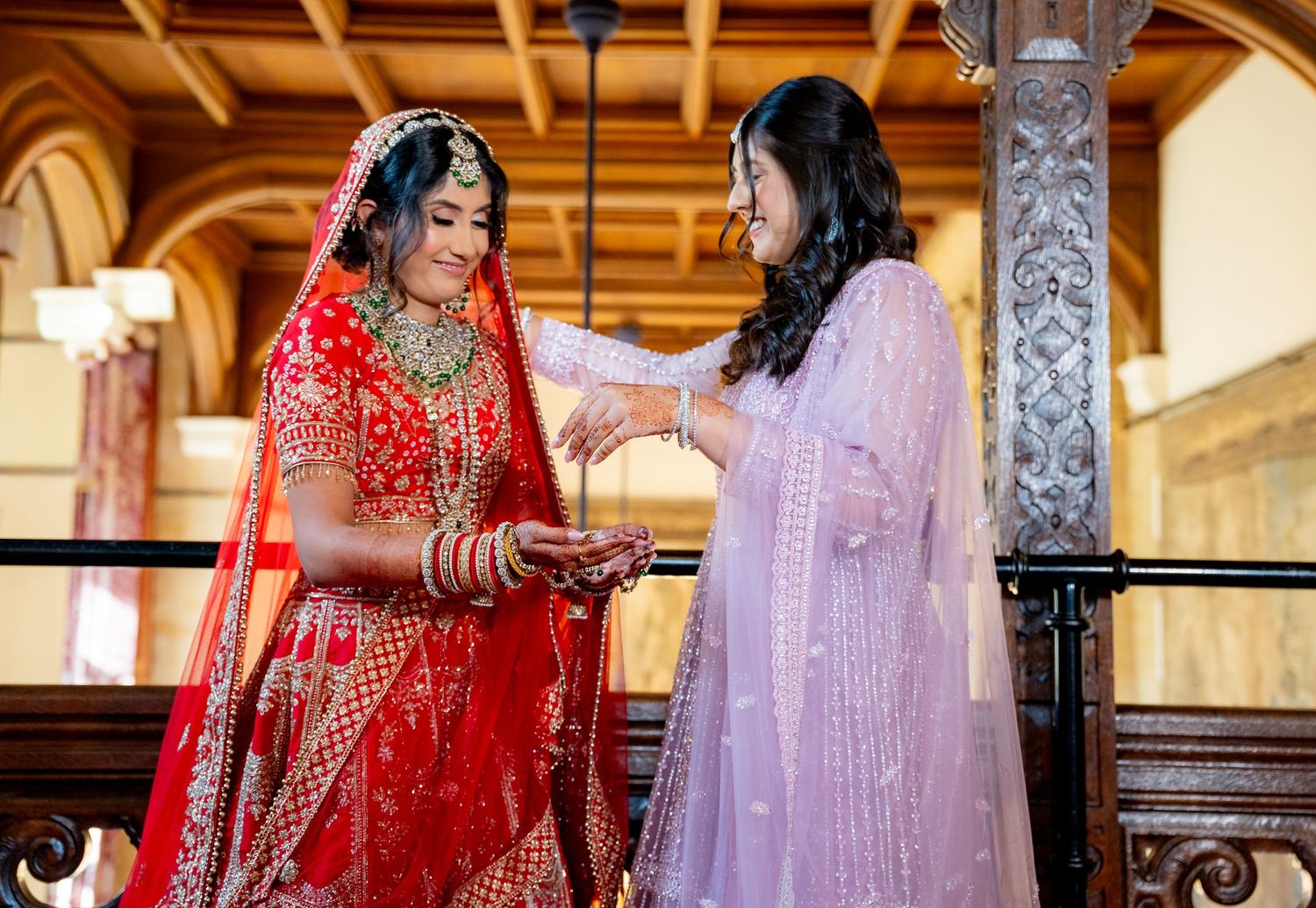 Indian bride in red lehenga receiving blessings from woman in lavender dress on ornate wooden balcony.
