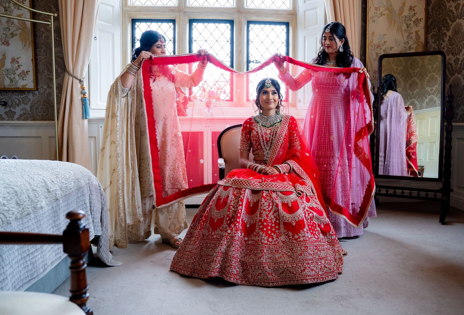 Indian bride in red lehenga has dupatta lifted by bridesmaids in floral wallpapered bridal suite.