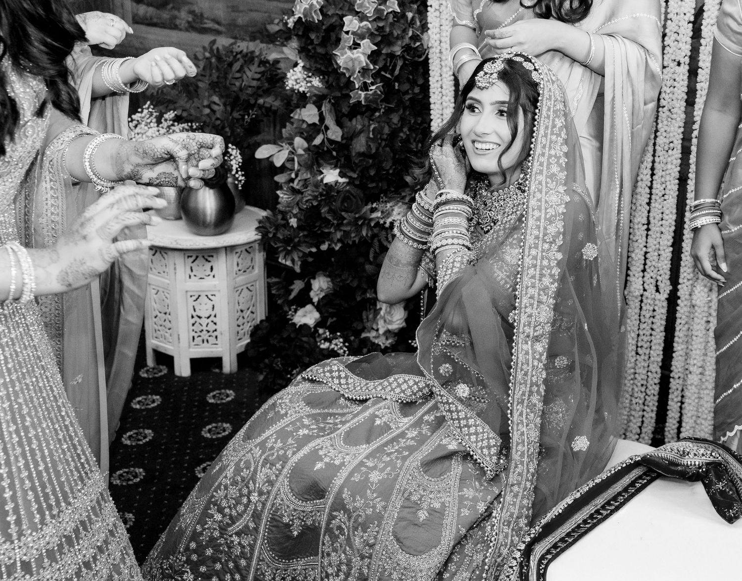 Black and white photo of Indian bride in embroidered lehenga smiling during traditional wedding ritual ceremony.