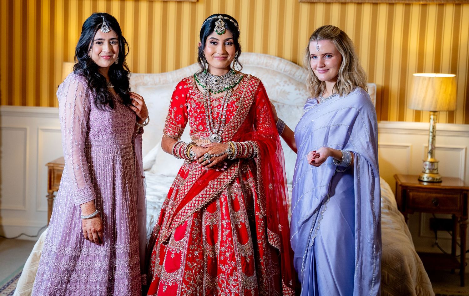 Indian bride in red lehenga poses with two bridesmaids in lavender and blue outfits.
