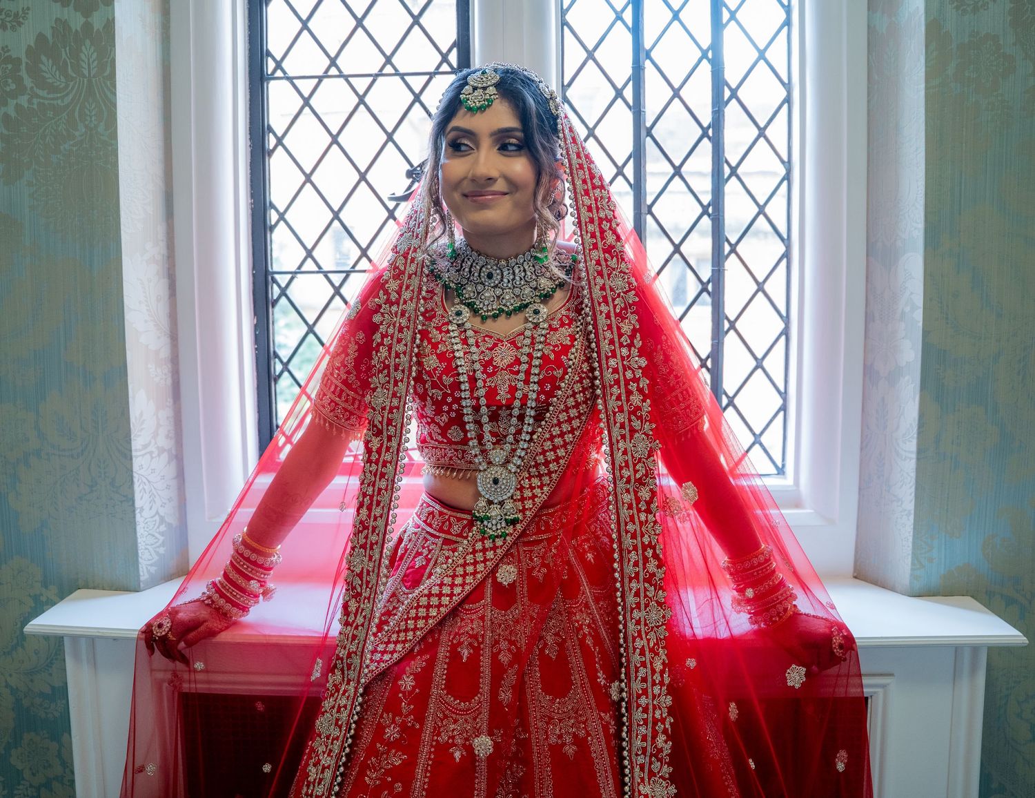 Indian bride in red lehenga choli standing by arched window in stunning traditional bridal ensemble.