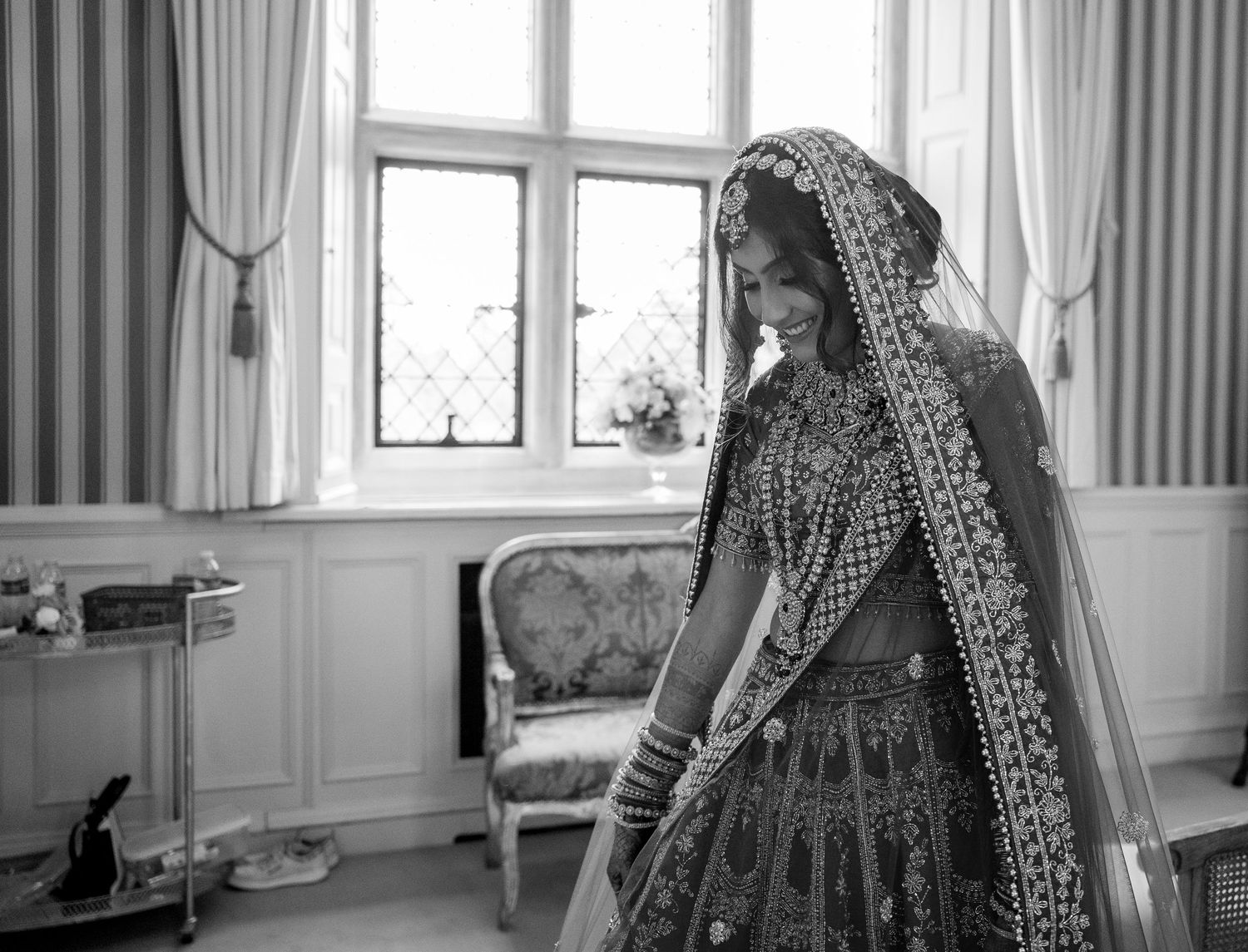 Black and white photo of a bride in traditional Indian bridal lehenga and dupatta standing by a window.