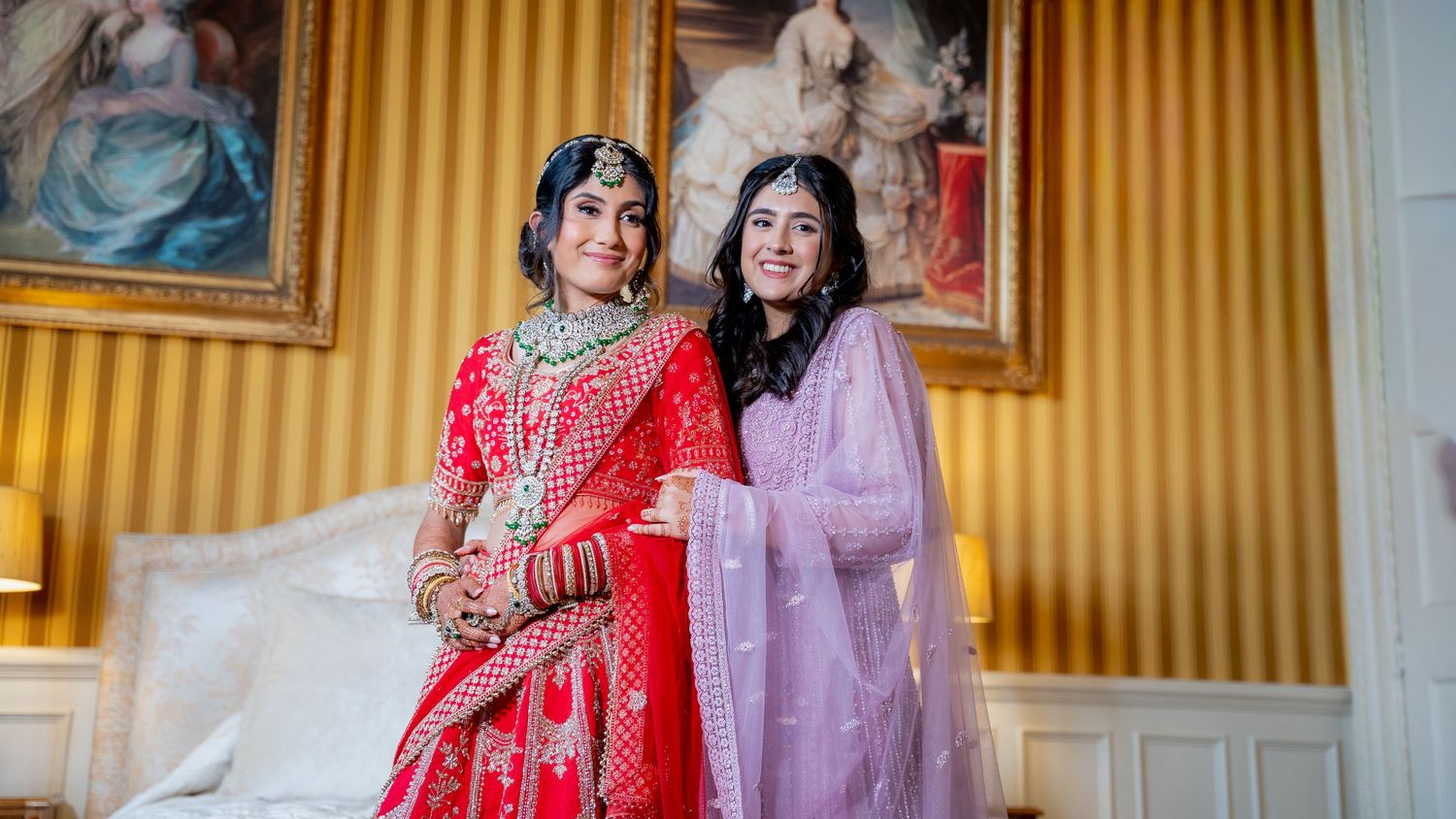 Bride in red bridal lehenga poses with guest in lavender salwar kameez in elegant bedroom.