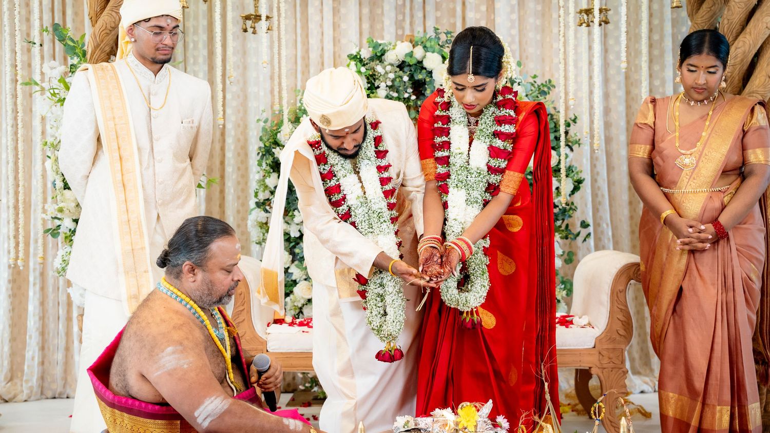 Indian bride in red saree and groom exchange vows during traditional Hindu wedding ceremony.