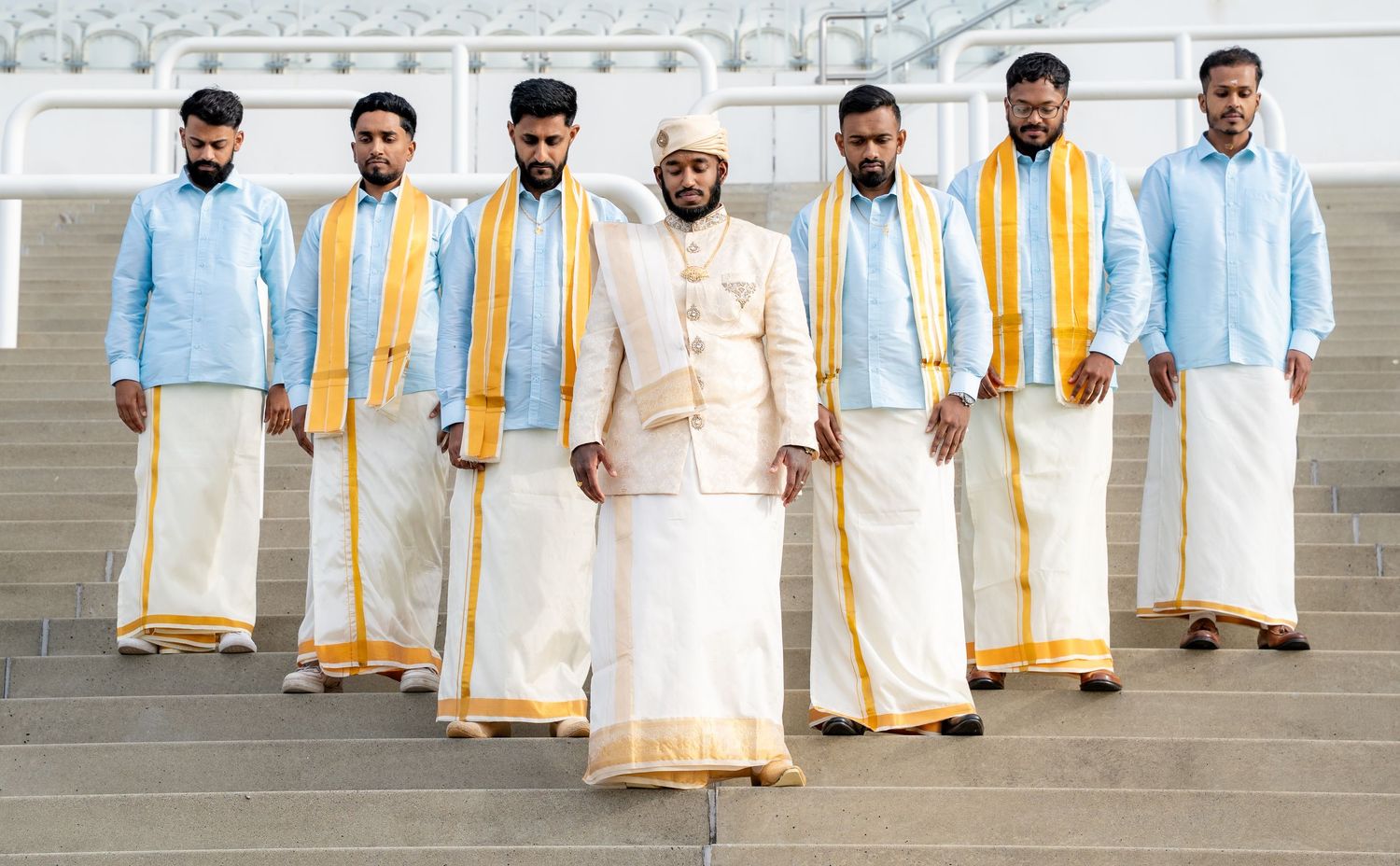 A groom in white and gold traditional attire stands with groomsmen in light blue shirts and white dhotis on steps.