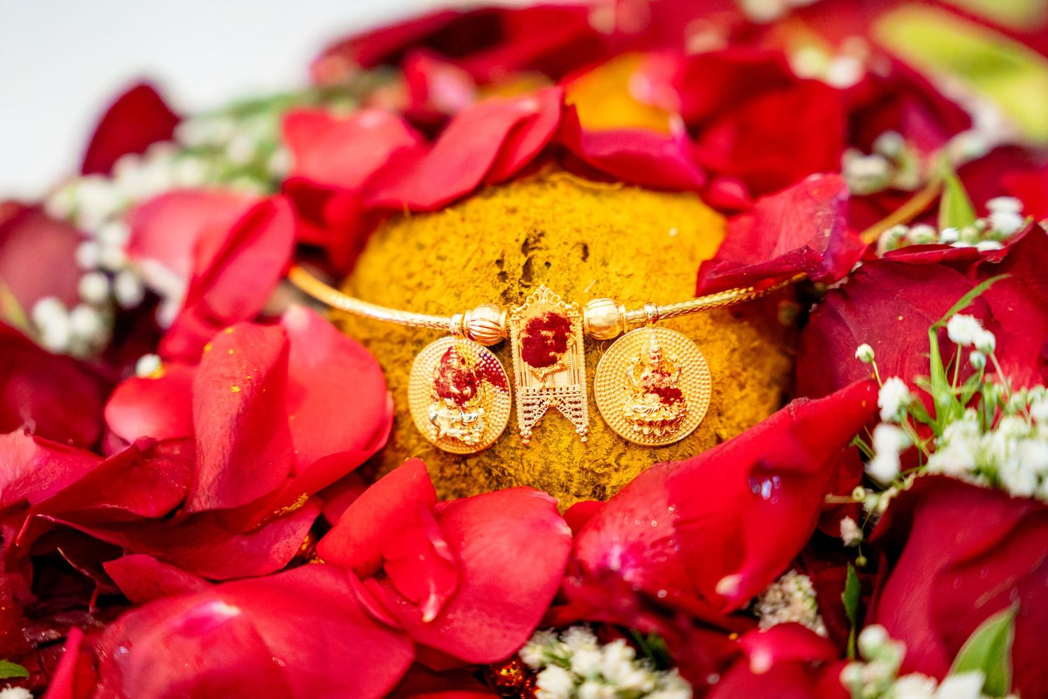 Ornate gold bangle with deity charms resting on turmeric powder amid vibrant red rose petals and white flowers.