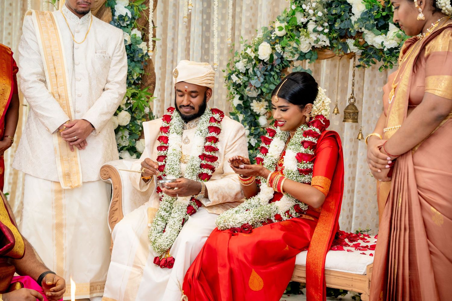 Couple seated together during traditional South Indian wedding ceremony with floral garlands and red bridal saree.