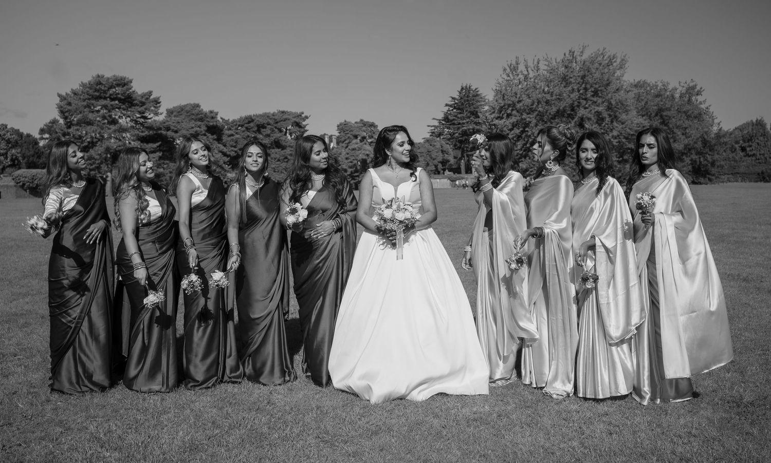 Black and white photo of bride in white gown posing with bridesmaids in elegant dresses outdoors.