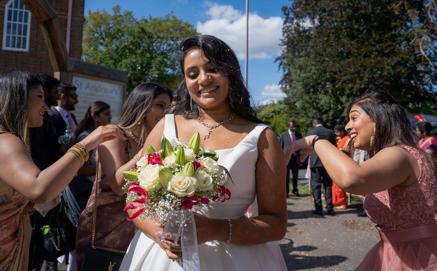Smiling bride in white dress holding bouquet of white and red flowers surrounded by guests outdoors on sunny day.