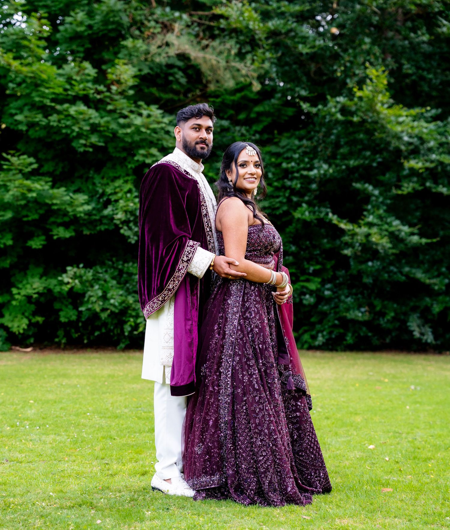 A couple poses on a green lawn, wearing matching deep purple Indian wedding attire.