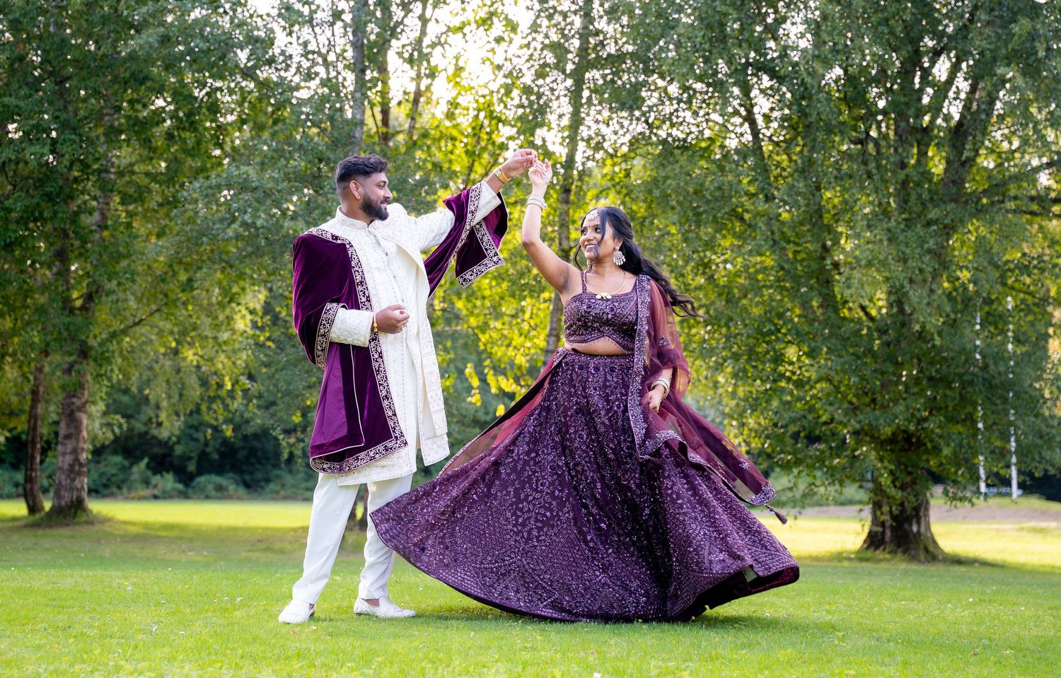 Couple in traditional Indian attire dancing outdoors, woman in purple lehenga twirling, man in white sherwani, lush green trees.