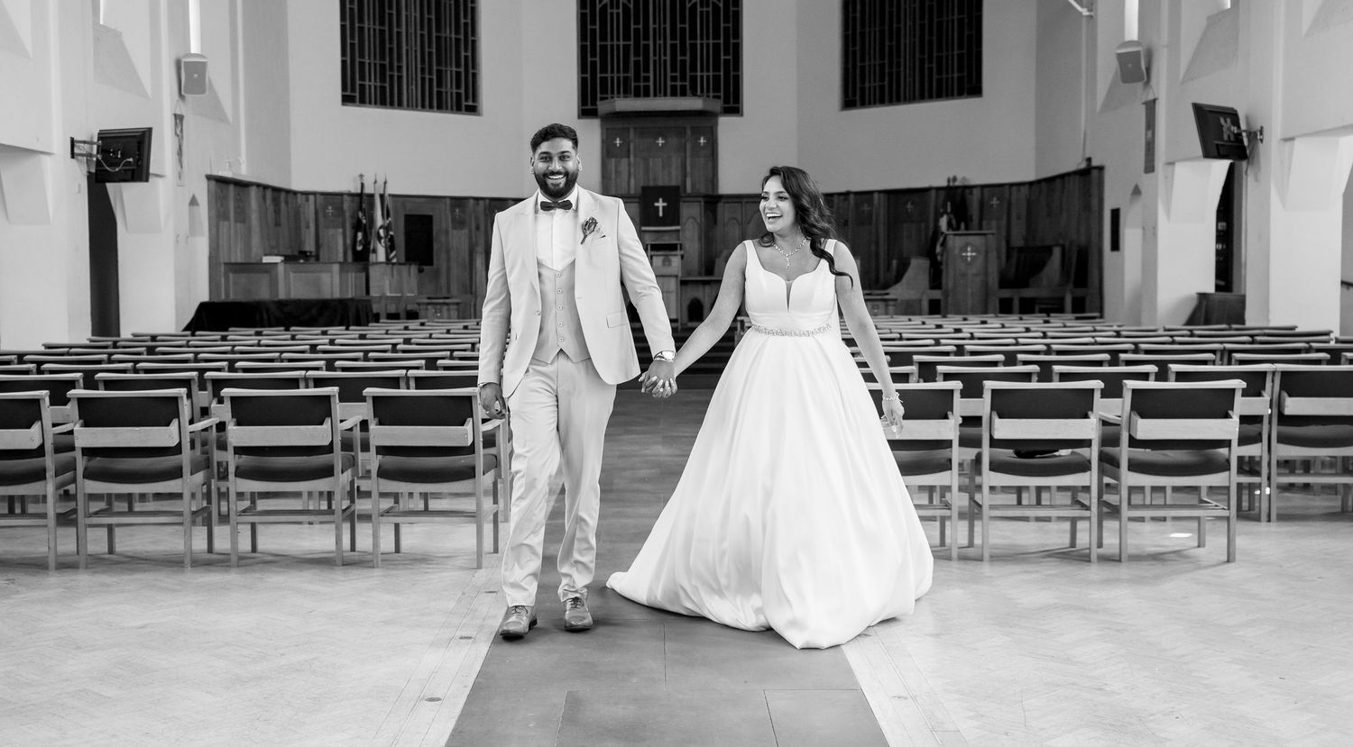 Black and white photo of bride and groom holding hands walking down the aisle of an elegant church.