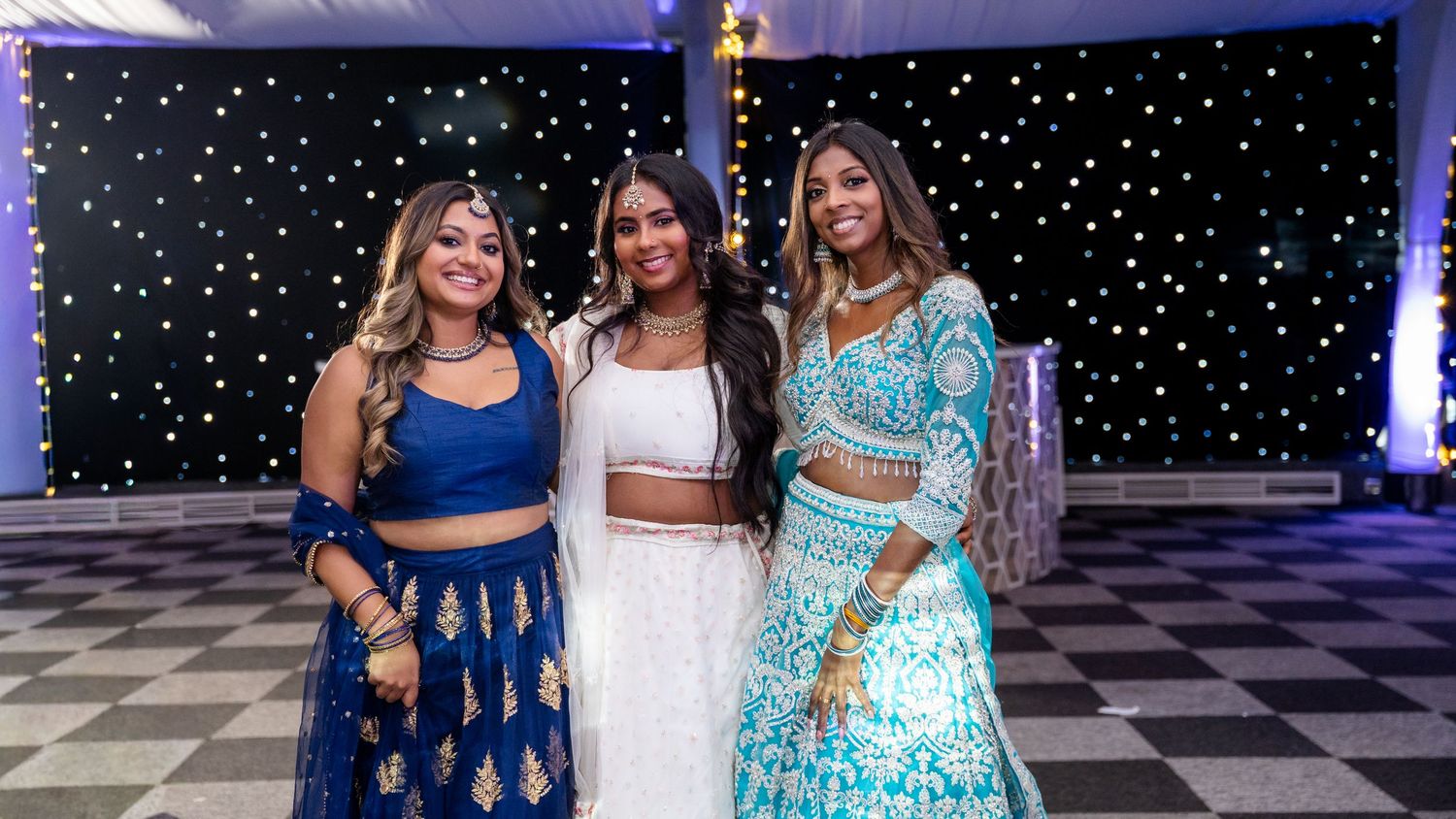 Three women posing in stunning blue and white Indian lehengas against a starlit black backdrop.