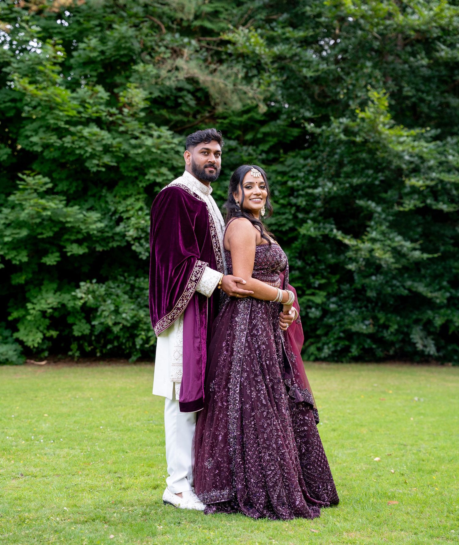 Couple in matching deep purple traditional South Asian attire posing outdoors on green lawn with lush trees behind them.