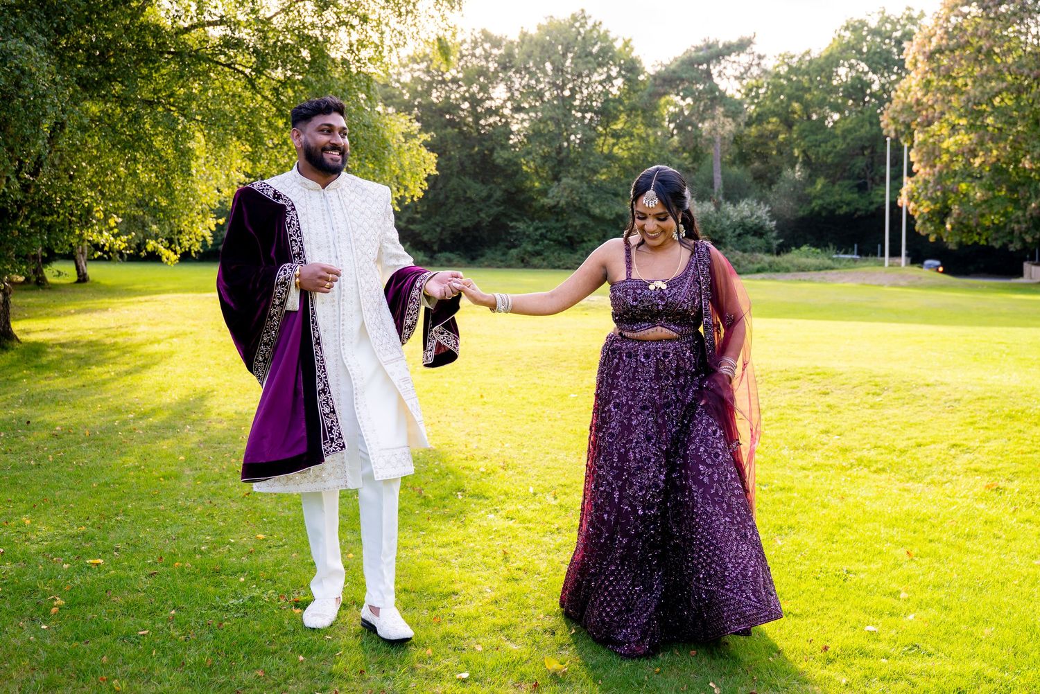 Indian couple in traditional purple and white wedding attire walking on green lawn with trees in background.