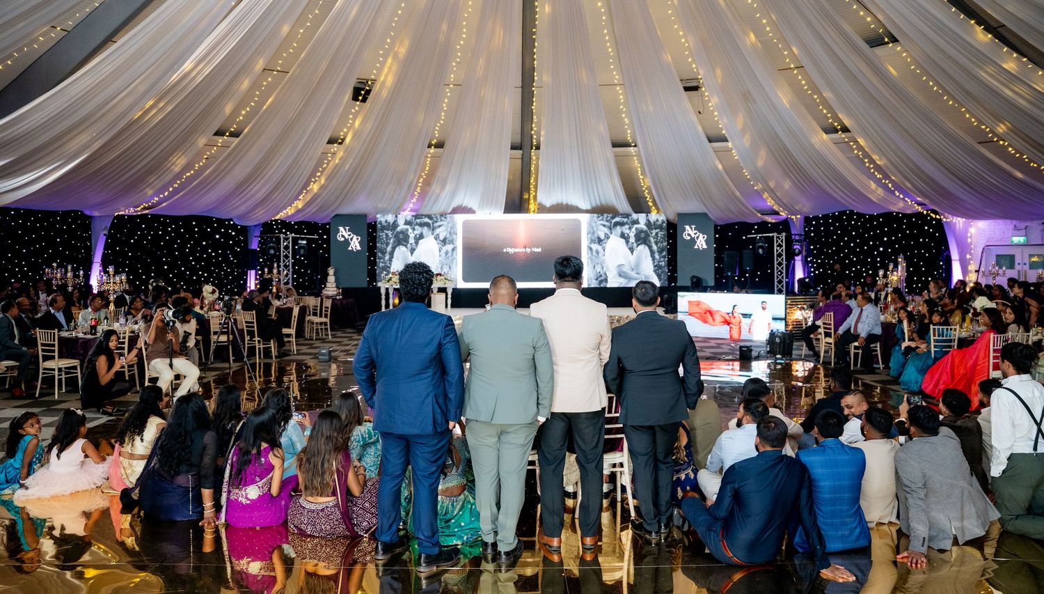 Elegant wedding reception in a decorated tent with draped ceilings, guests gathered around a dance floor with a large screen.