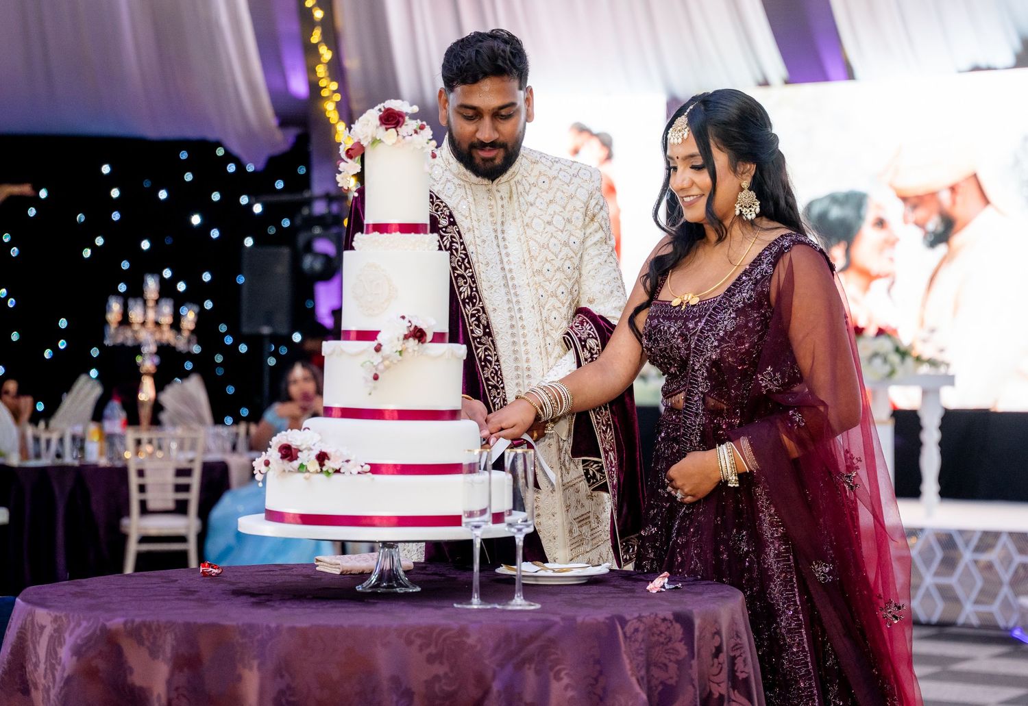Bride and groom in traditional South Asian attire cutting a white four-tier wedding cake at an elegant reception.