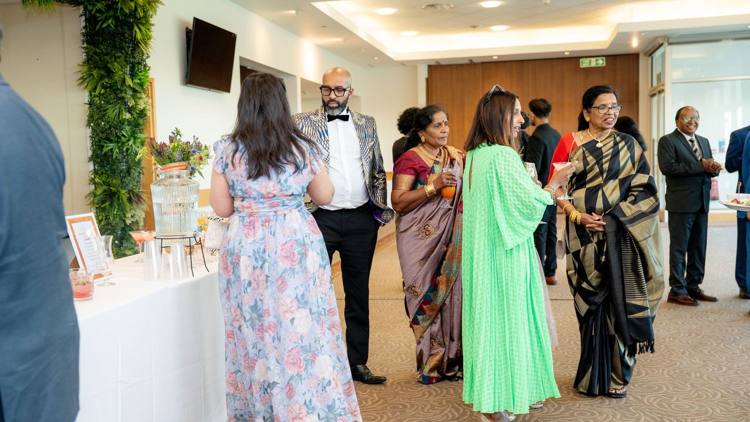 Elegantly dressed wedding guests socializing in a bright venue with green floral decorations.