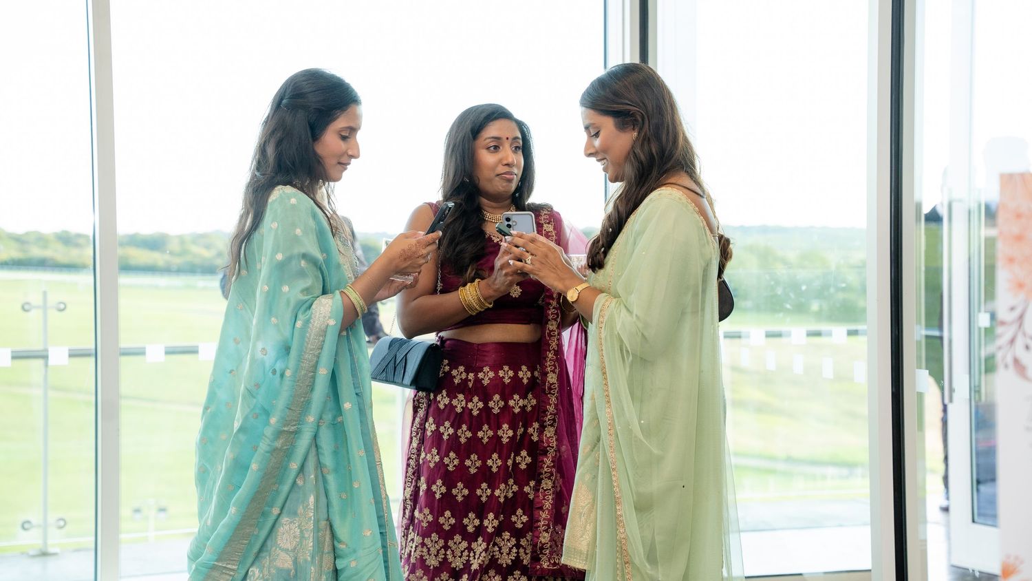 Three women in Indian traditional sarees conversing by large windows at a wedding reception.