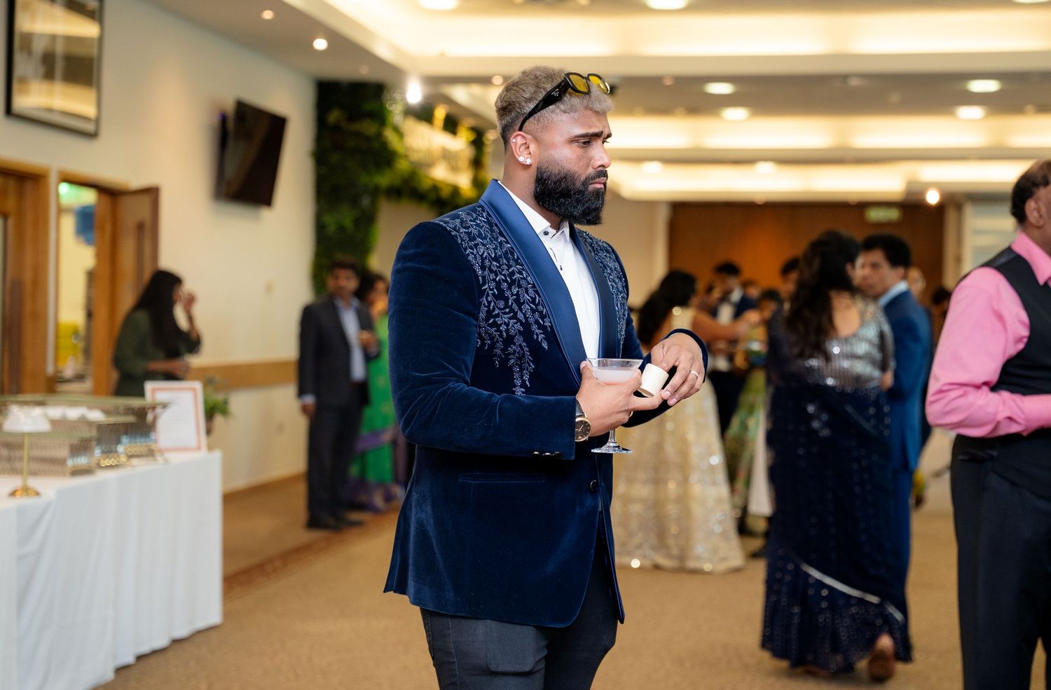 Man in navy blue velvet blazer holding a drink at an elegant indoor social event with guests in formal attire.