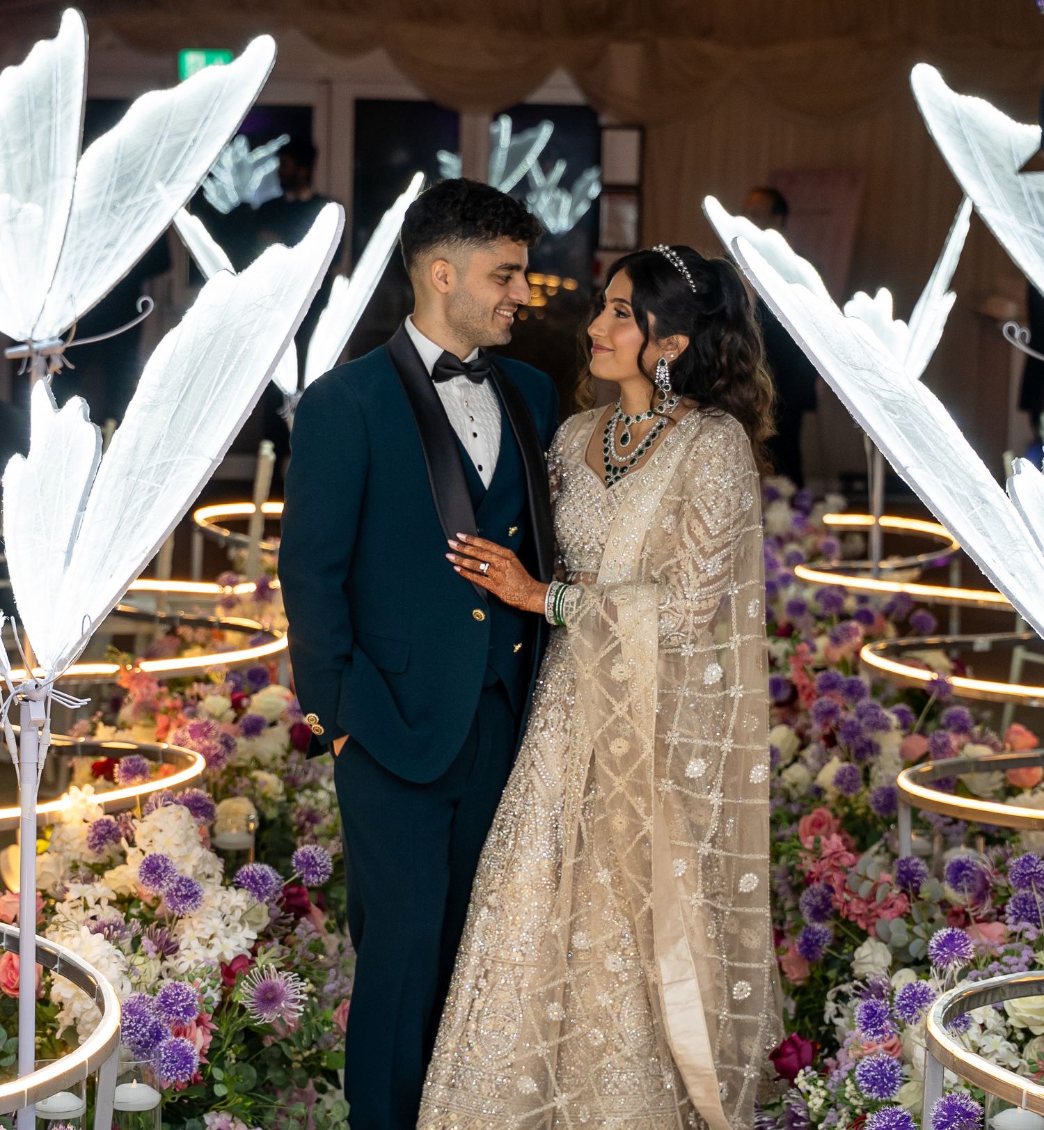 Couple in elegant wedding attire share romantic moment surrounded by floral arrangements and butterfly lights.