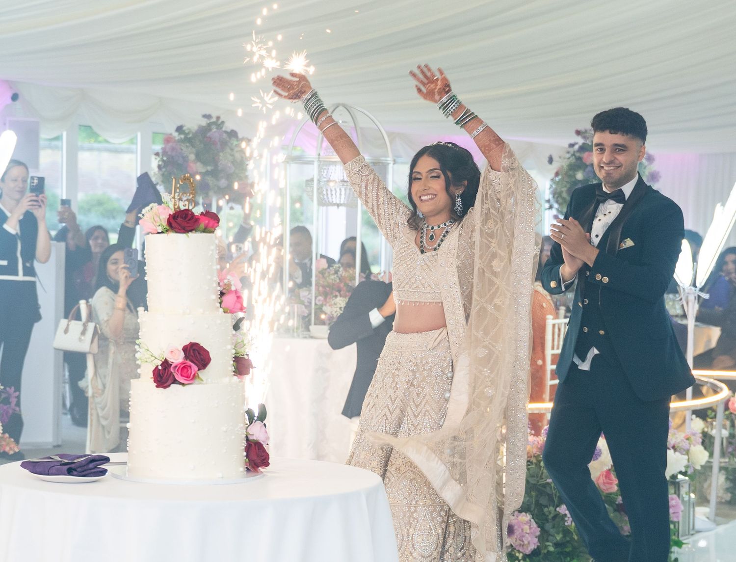 South Asian bride and groom celebrate wedding cake cutting with sparklers in elegant white marquee venue.