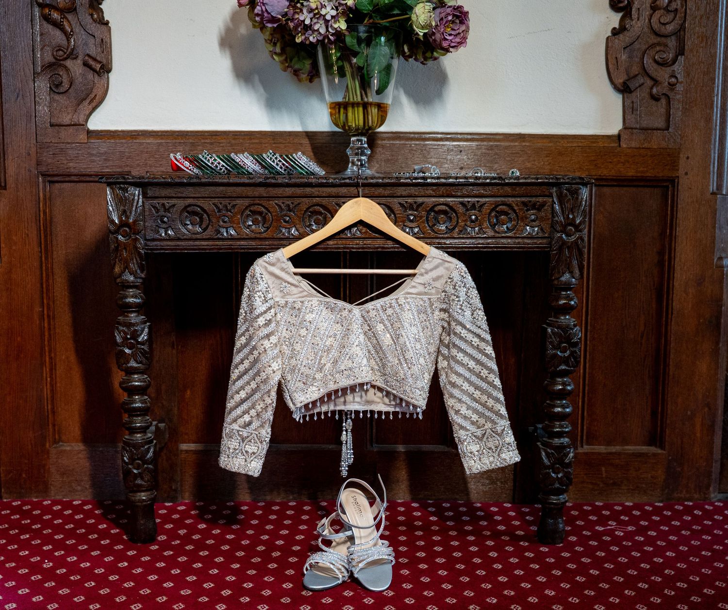 Ivory embroidered blouse hanging on hanger above blue heeled sandals on ornate wooden console table.