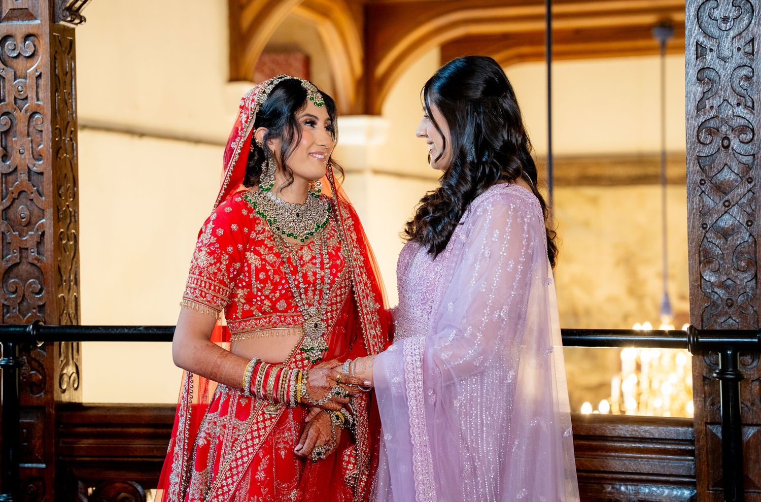 Indian bride in red lehenga sharing a moment with bridesmaid in pink.