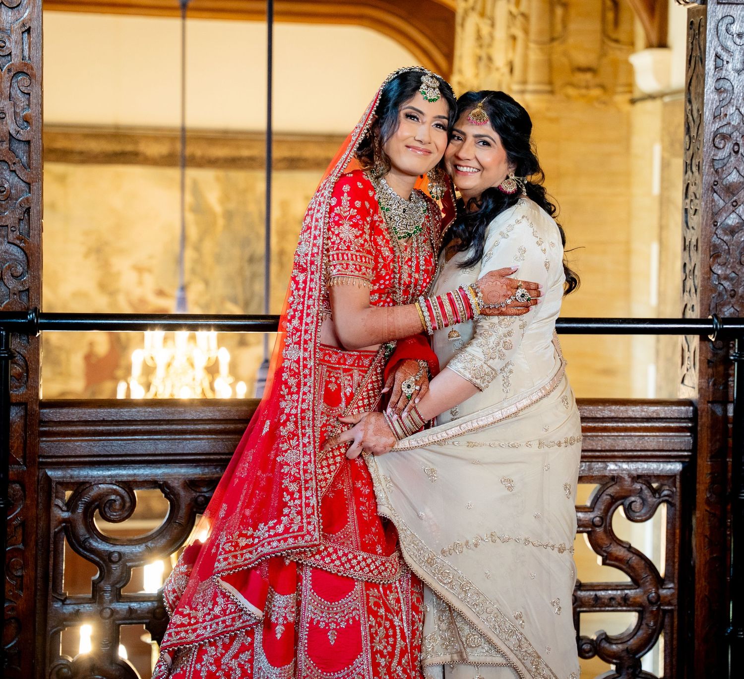Indian bride in red lehenga poses with woman in cream saree at ornate wedding venue.