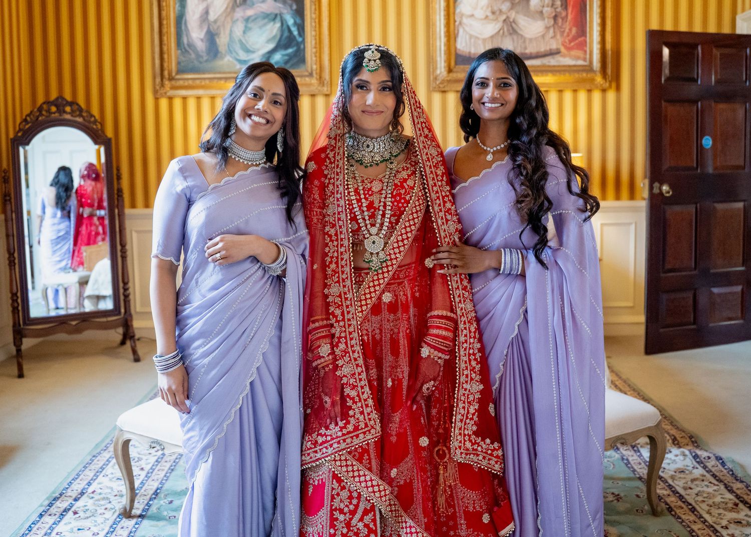 Indian bride in red lehenga posing with two bridesmaids in lavender sarees in ornate room.