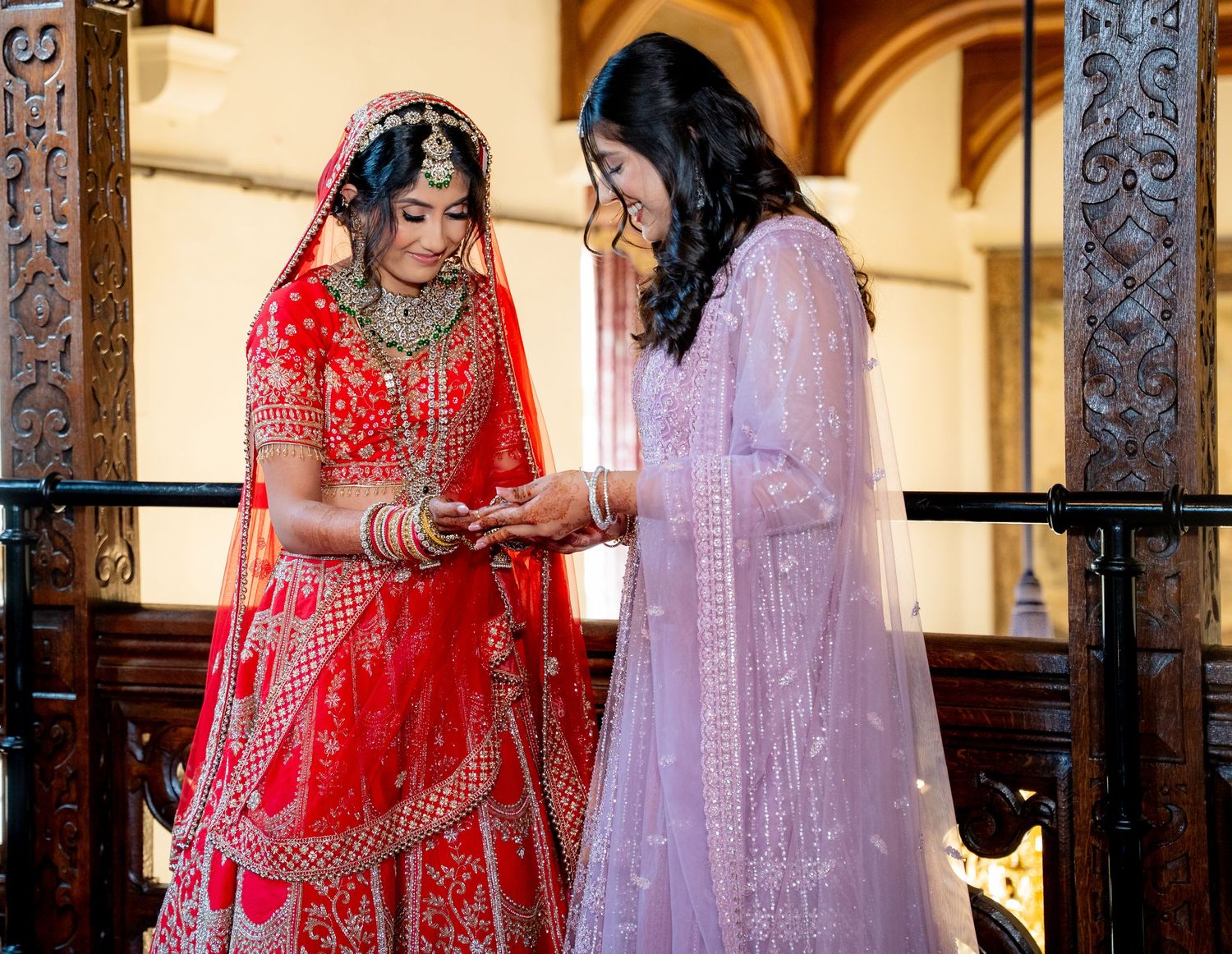 Indian bride in red bridal lehenga and woman in lavender salwar examining jewelry on wooden balcony.