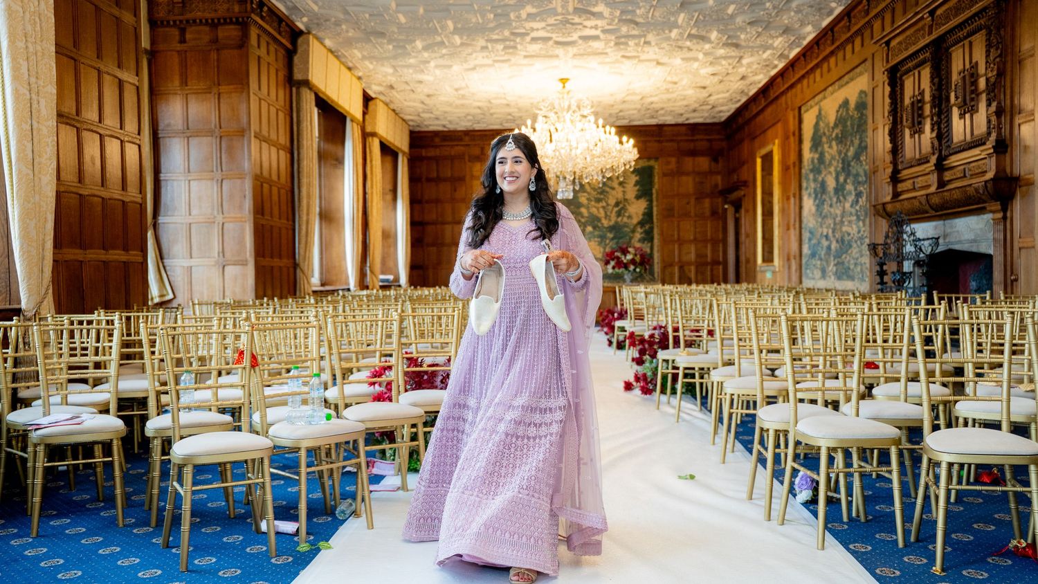 Bride in lavender lehenga poses in ornate ballroom decorated with gold chairs for Indian wedding.