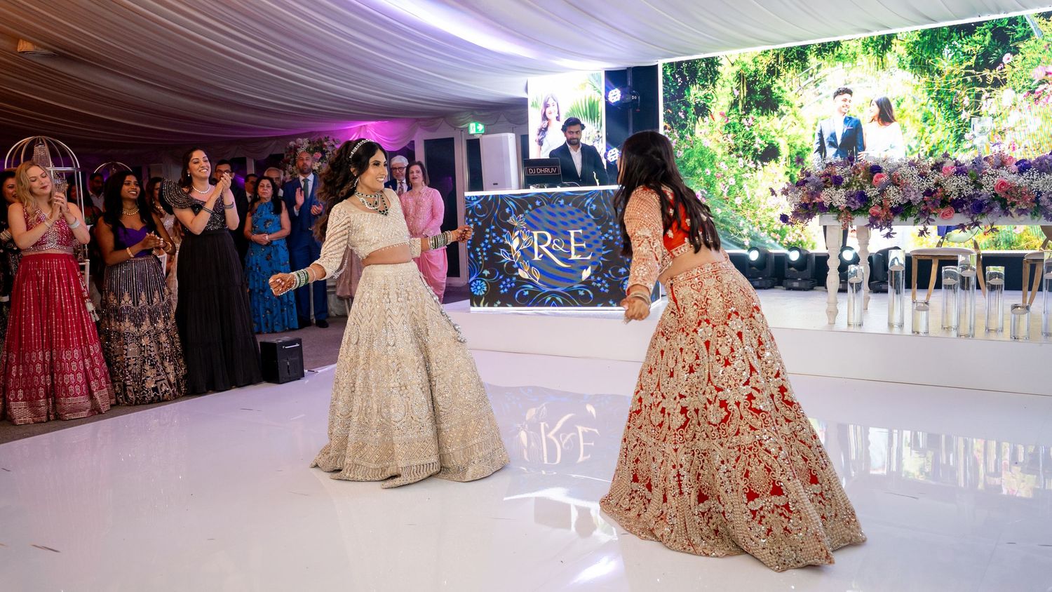 Two women in elegant Indian lehengas perform a dance at a wedding reception on a white dance floor.