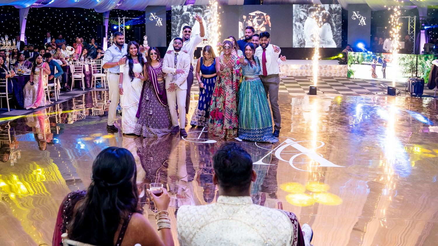 Wedding party group photo on dance floor with fountains and guests at Indian wedding reception.