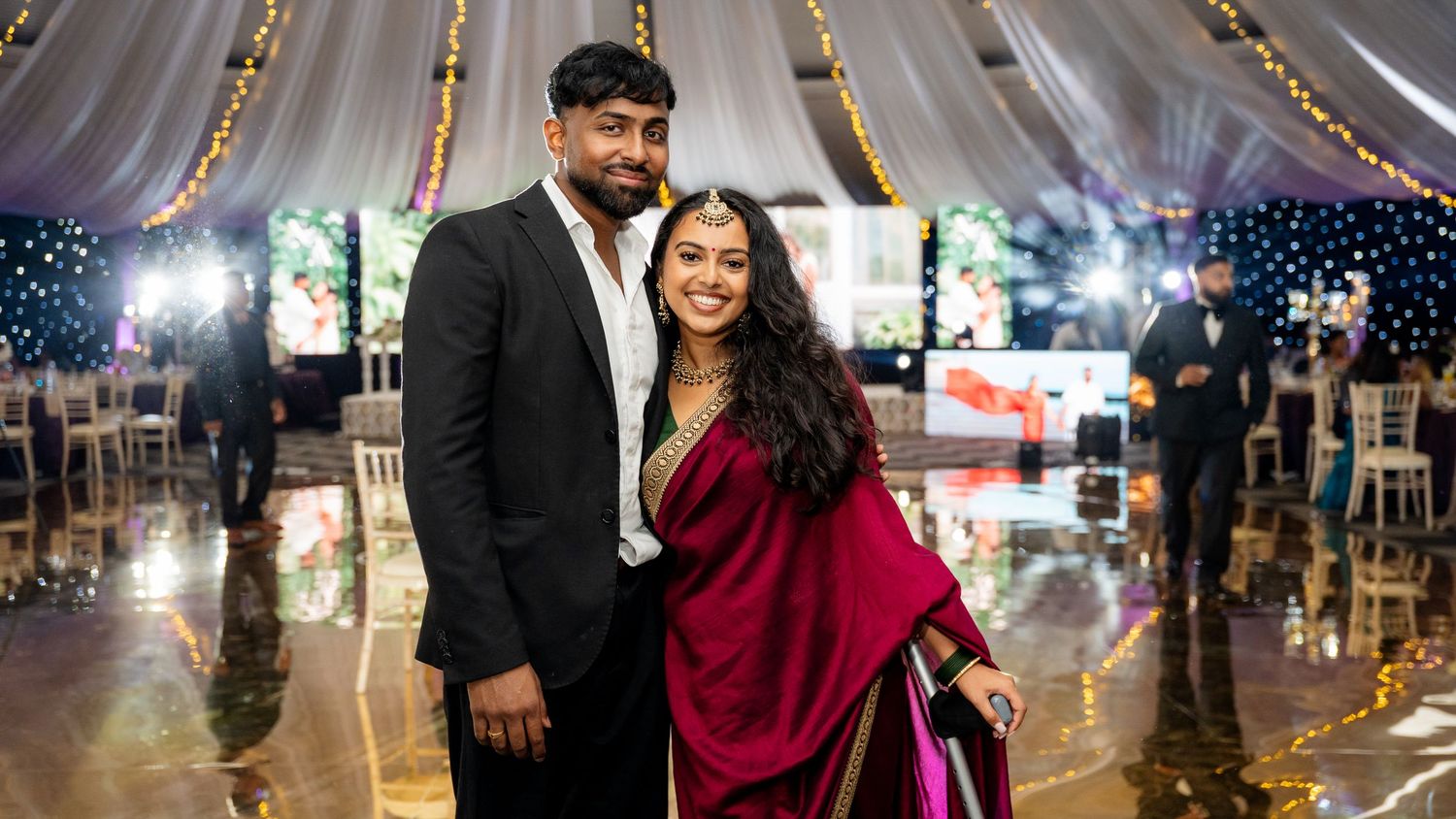 Couple posing together smiling in elegant Indian wedding attire inside decorated reception venue.