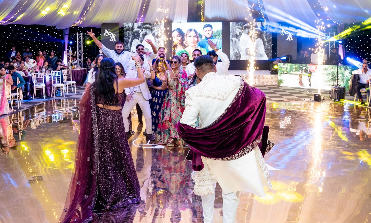 Bride in purple lehenga and groom in white sherwani dancing joyfully at Indian wedding reception.