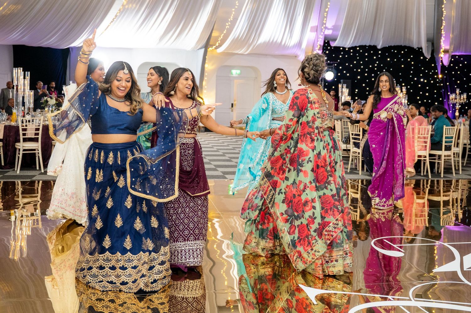 Ladies in colorful Indian lehengas and sarees dancing together at a festive wedding reception.