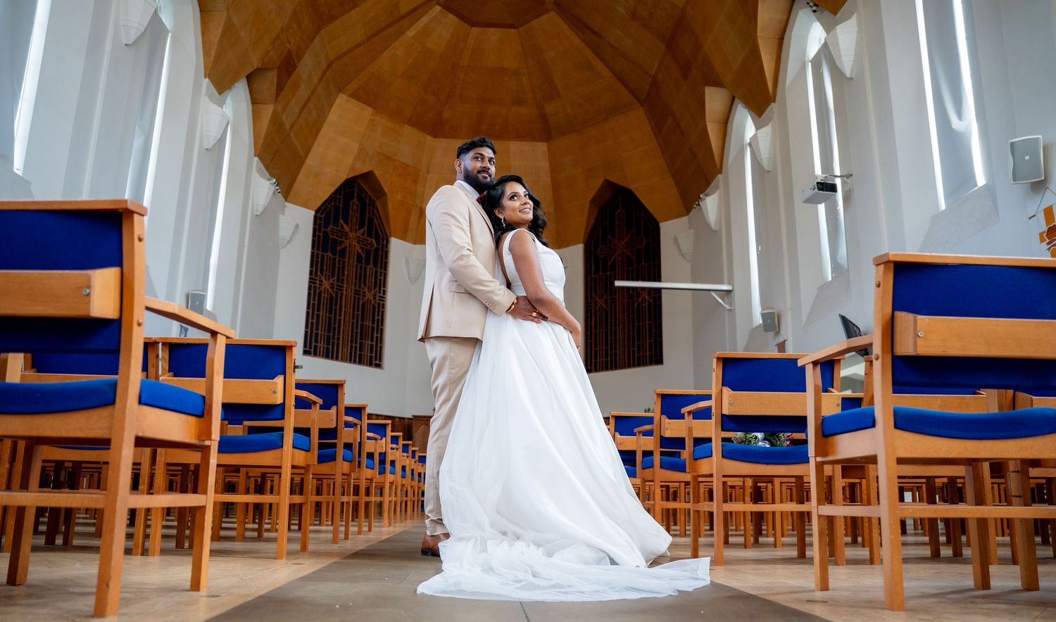 Bride and groom posing elegantly in a church aisle surrounded by blue chairs and wooden arched ceiling.