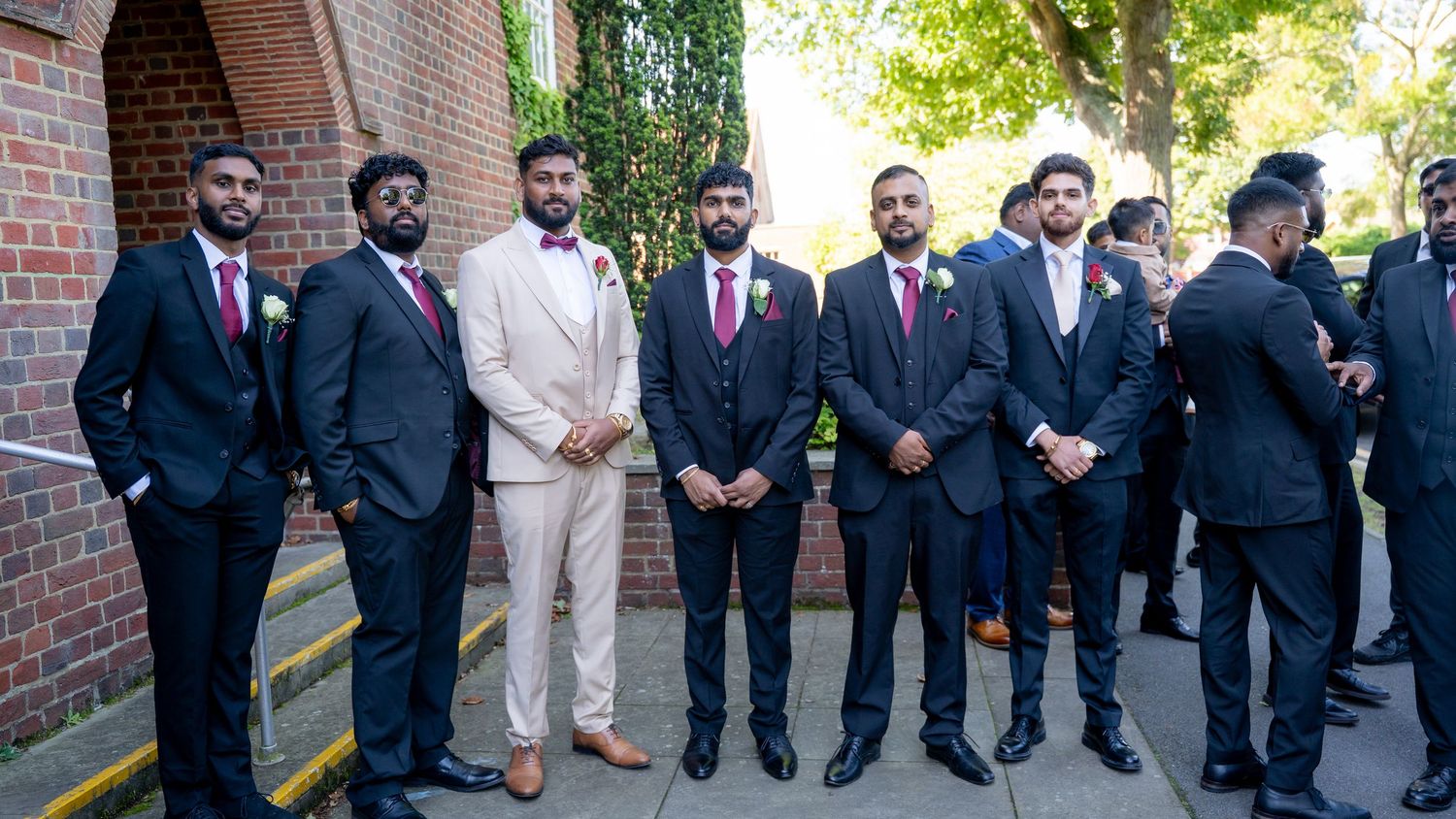 Groomsmen in navy suits with floral boutonnieres stand outdoors near a brick wall at a wedding ceremony.