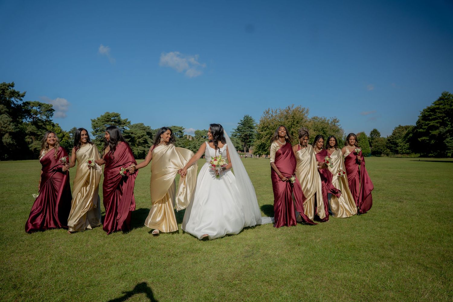 Bride in white wedding gown walks with bridesmaids in red and gold sarees across green field.
