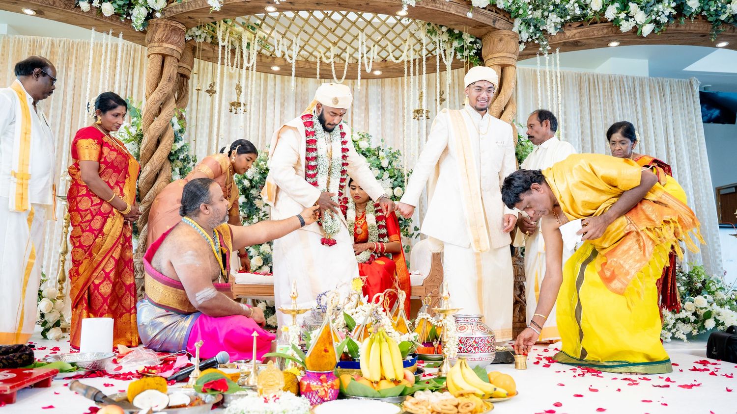 Traditional South Indian wedding ceremony with priest, bride in red saree, and groom in white dhoti exchanging garlands on decorated mandap.