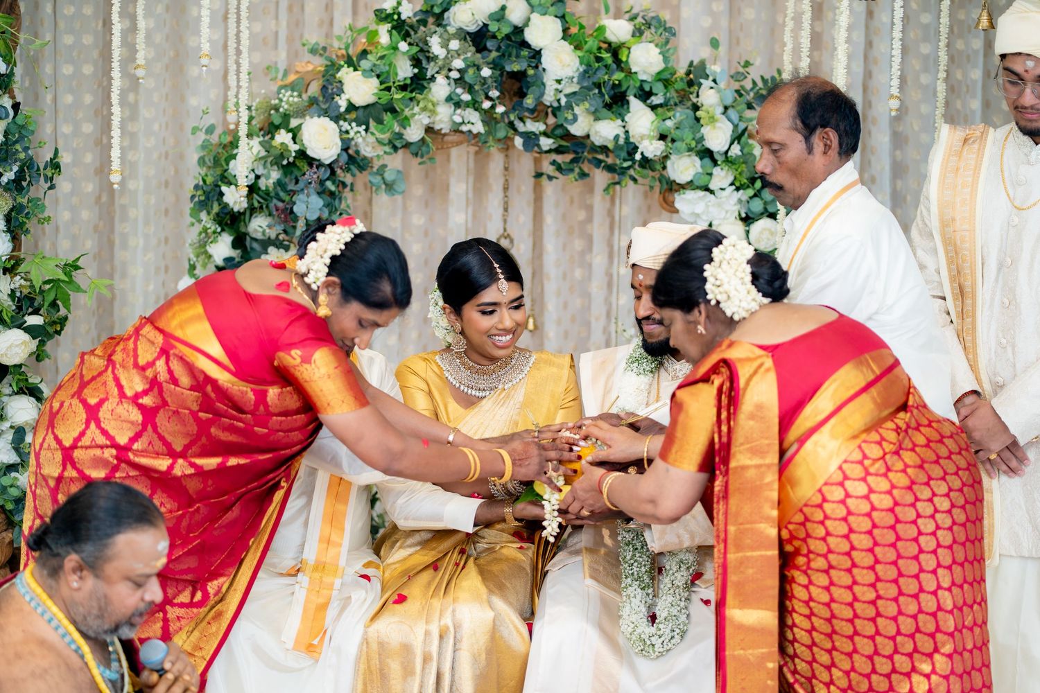Tamil wedding ceremony with women in red silk sarees performing traditional rituals for bride and groom.