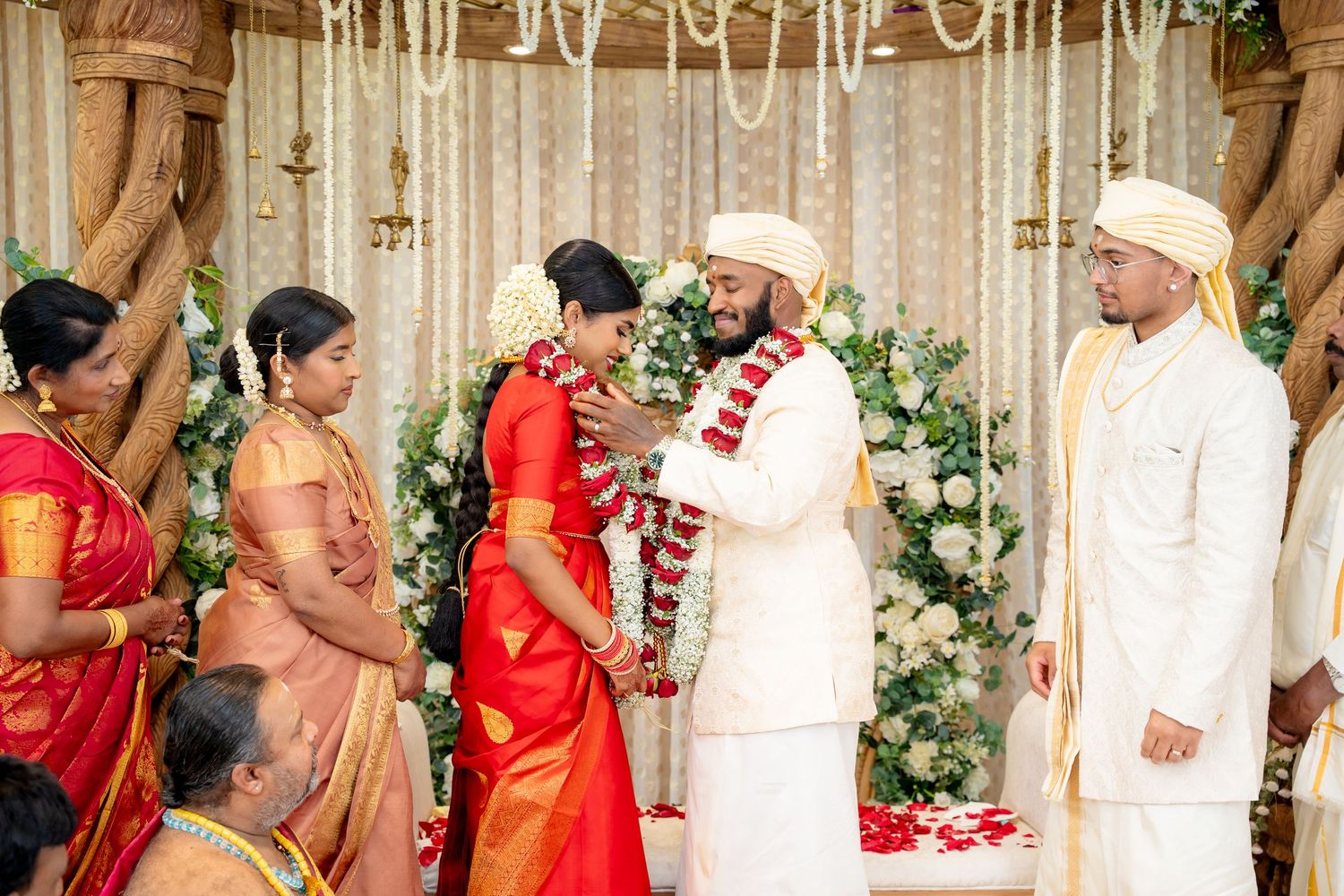Groom smiles at bride during traditional South Indian wedding garland exchange on an ornate mandap stage.