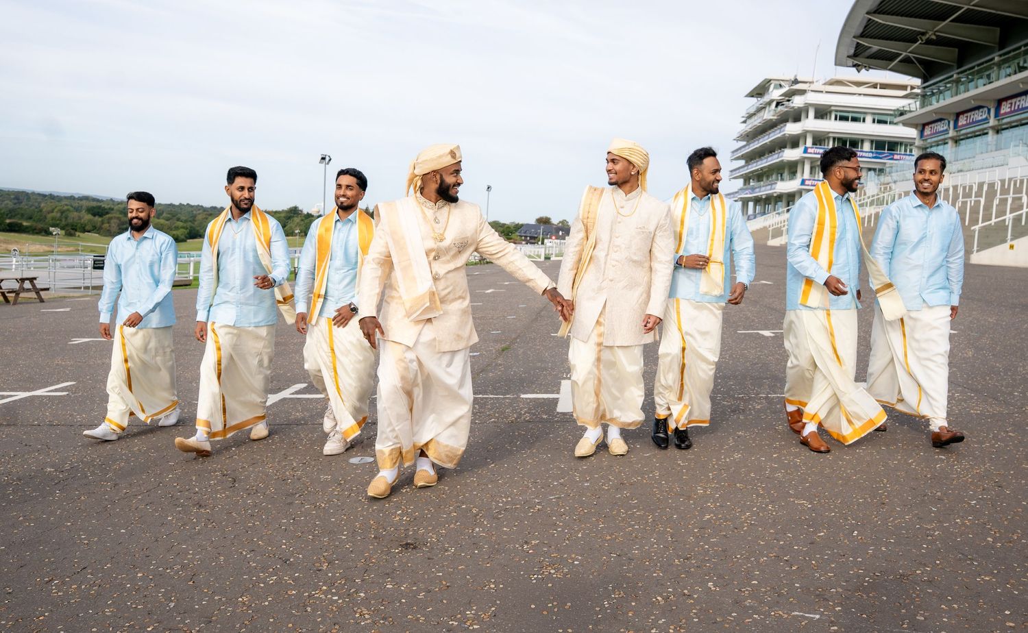 Group of men in traditional white clothing and yellow scarves walking together at an outdoor location with buildings in background.