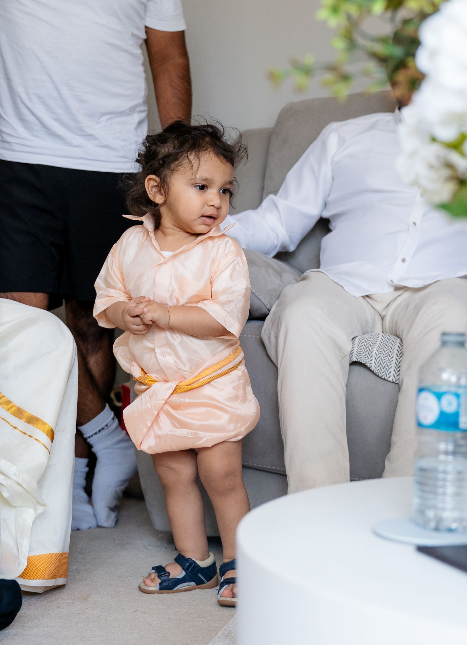 Adorable toddler in peach traditional outfit standing between adults at an Indian wedding celebration.