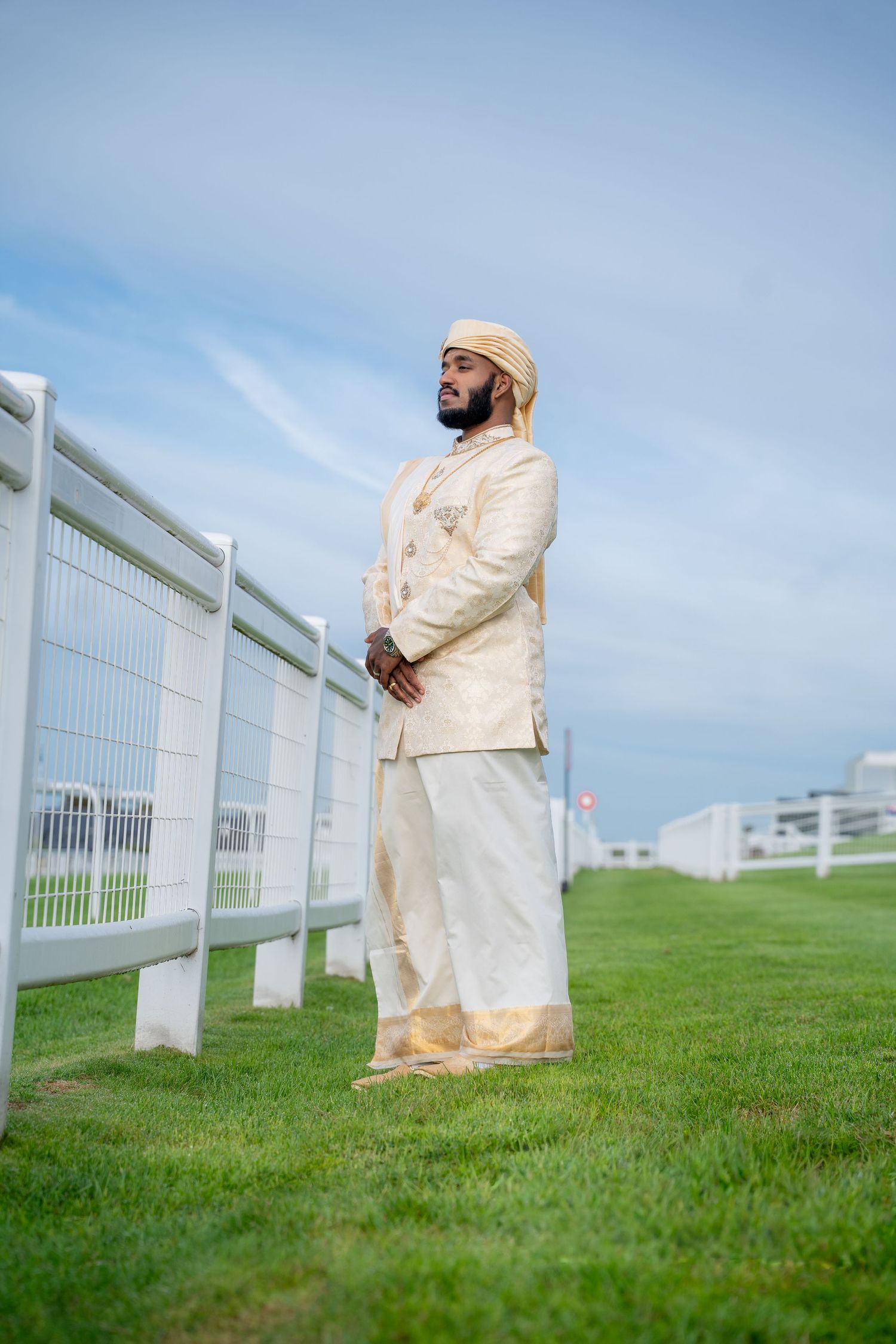 Groom wearing traditional cream gold sherwani posing beside white fence on green racecourse lawn.