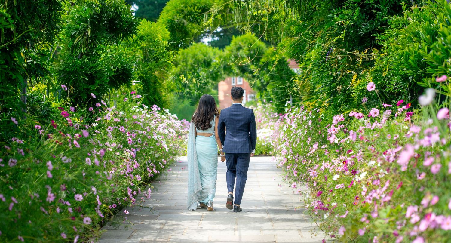 Couple walking hand in hand through flower-lined garden path in wedding attire.