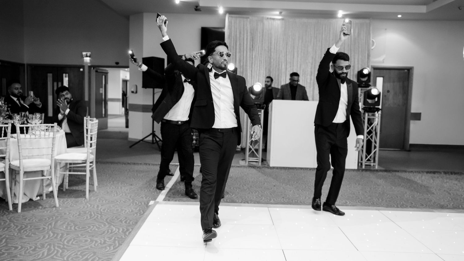 Black and white photo of groomsmen in tuxedos dancing with raised hands on a wedding dance floor.